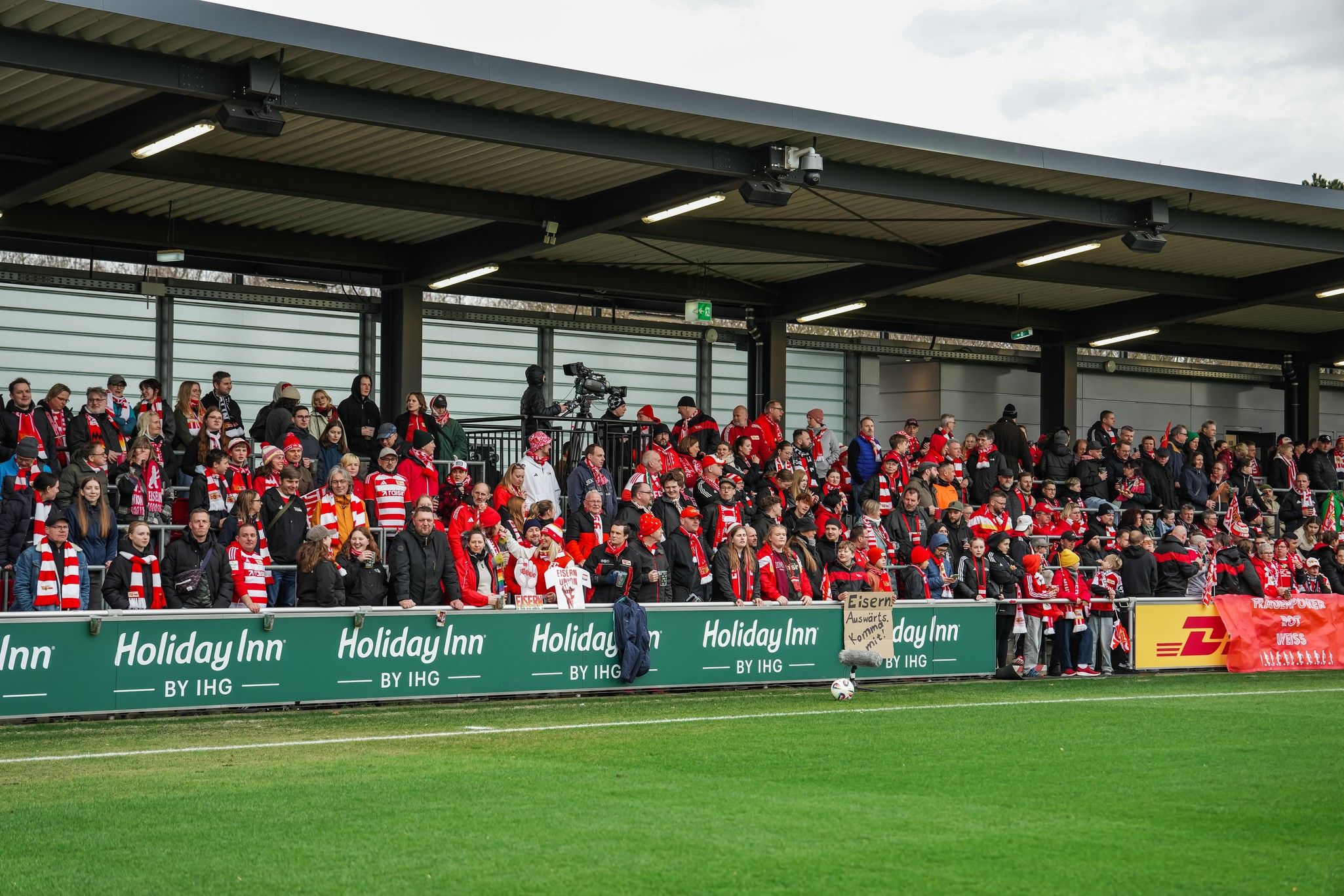 Zuschauer im Stadion feuern eine Fußballmannschaft an, umgeben von Werbebannern in rotem und schwarzem Design.