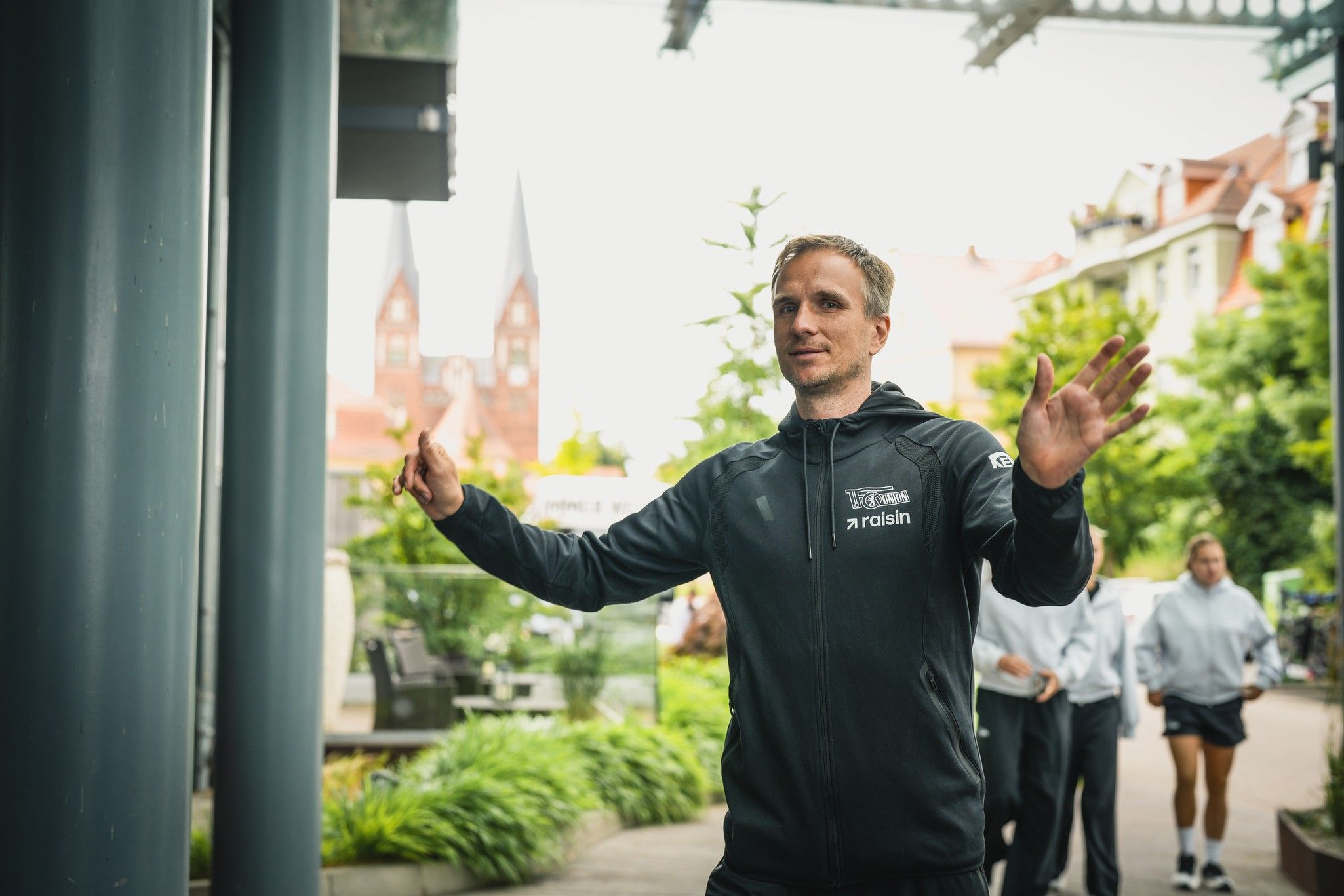Ein Mann in schwarzer Sportbekleidung steht im Freien mit erhobenen Händen. Im Hintergrund sind verschwommene Gebäude und ein Park zu sehen.