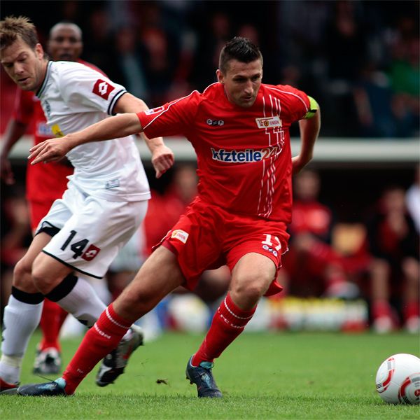 Ein Fußballspiel mit zwei Spielern: einer in rotem Trikot dribbelt, während der andere in weißem Trikot ihn verfolgt.