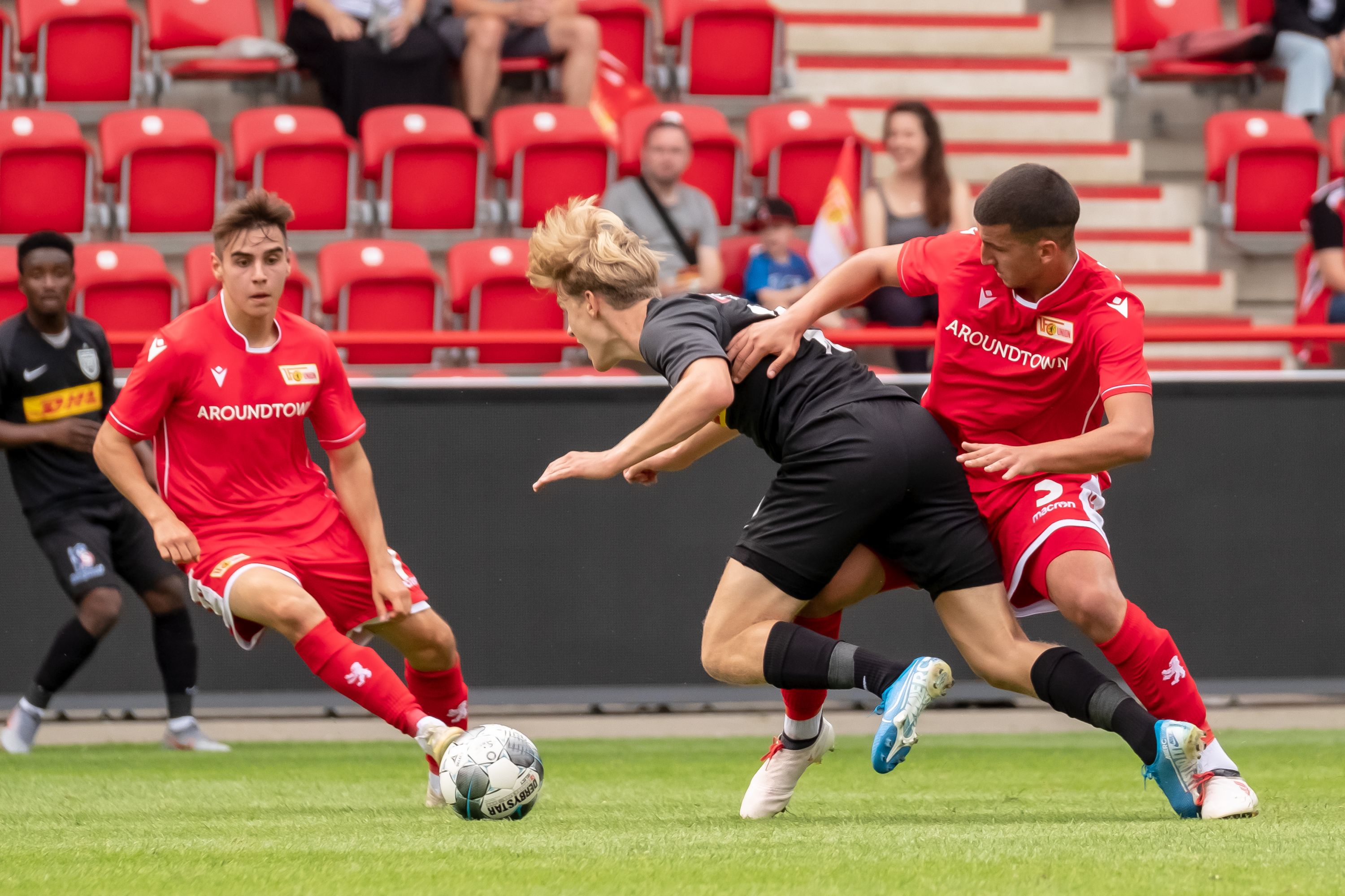 Two players in red jerseys are chasing a player in a black jersey during a soccer match on a stadium field.
