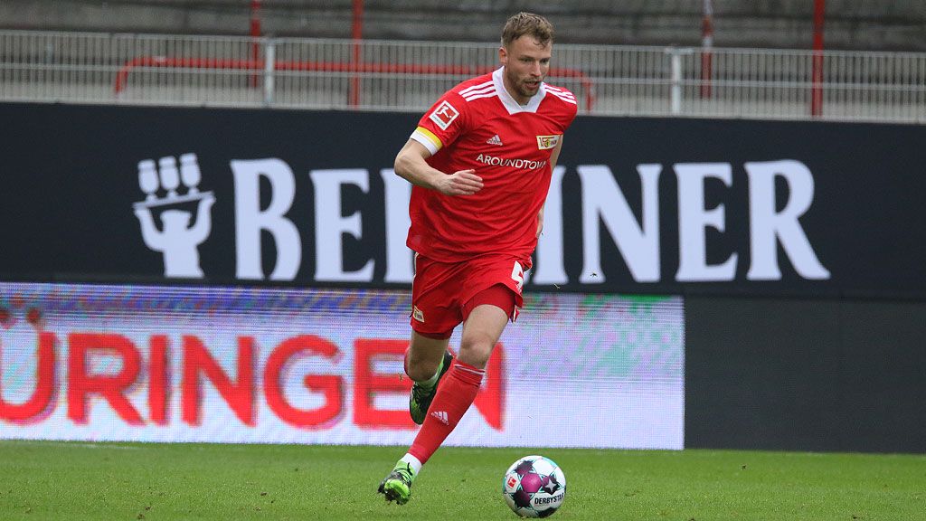 A soccer player in a red jersey is dribbling the ball on the field. Stadium stands can be seen in the background.