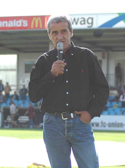 Ein Mann mit Mikrofon steht auf einem Sportplatz, im Hintergrund Zuschauer und Werbebanner sichtbar.