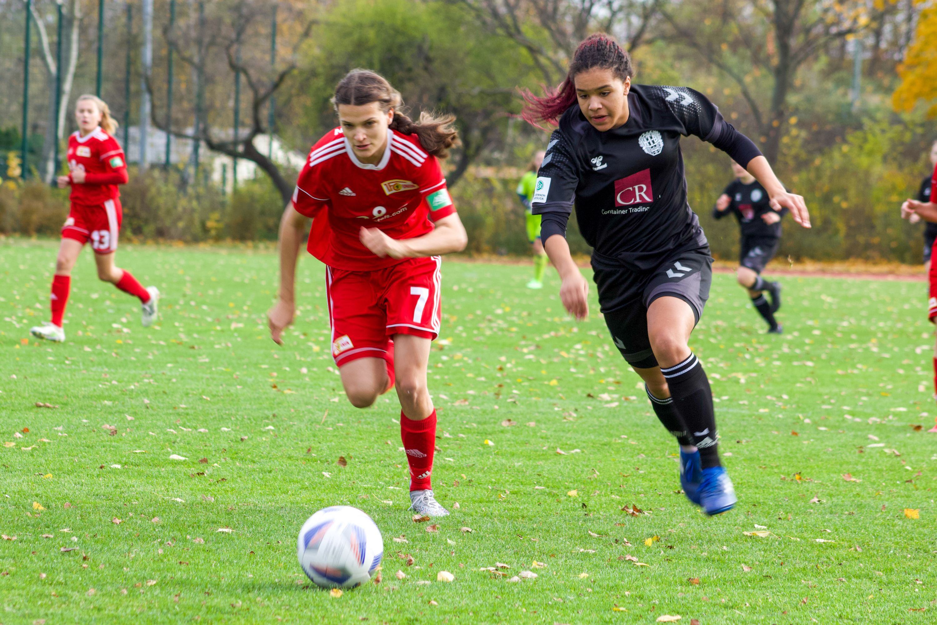 Zwei Fußballspielerinnen verfolgen den Ball auf einem grünen Spielfeld, umgeben von herbstlichen Bäumen.