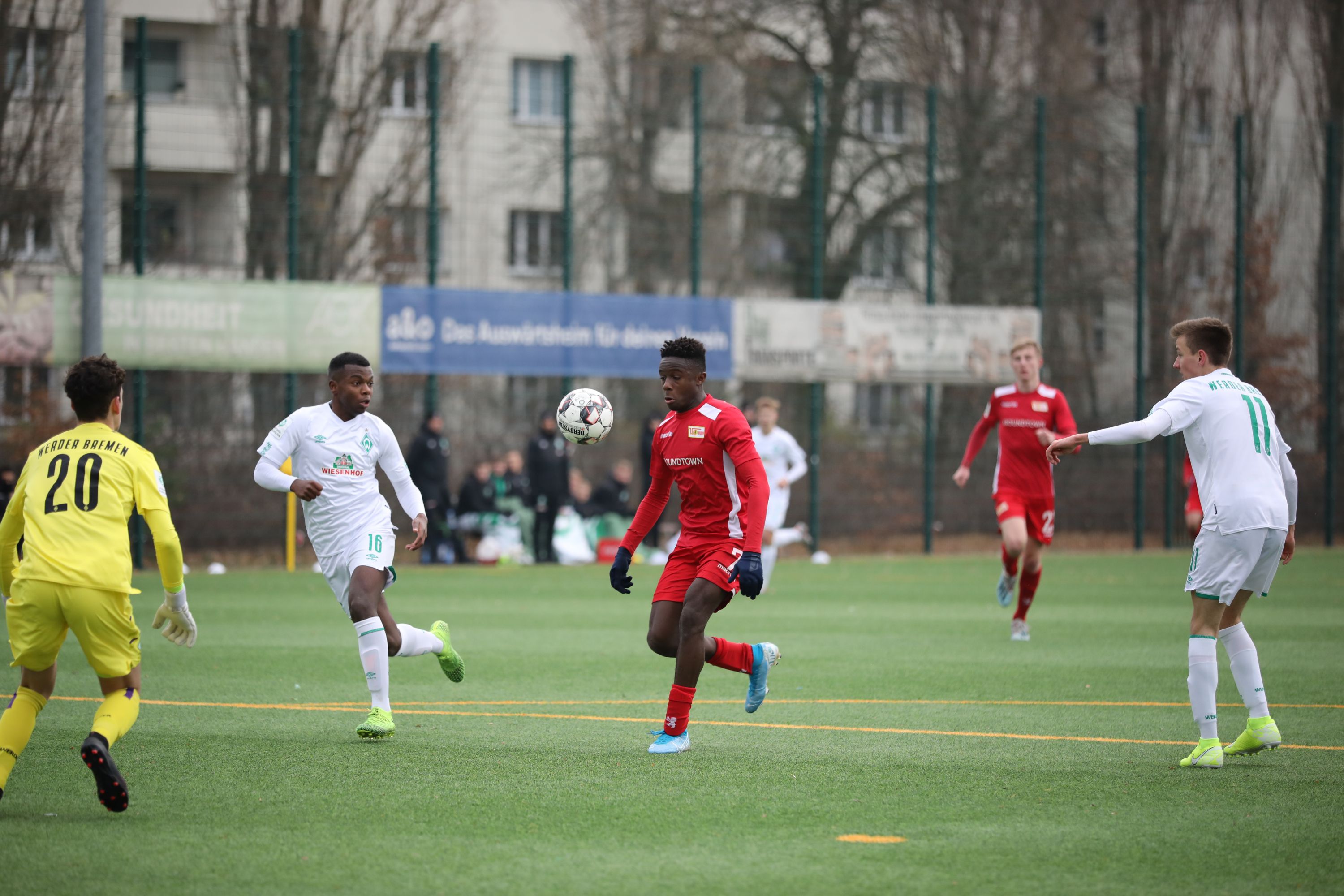 Spielszene eines Fußballspiels mit Spielern in roten und weißen Trikots auf einem Kunstrasenfeld.
