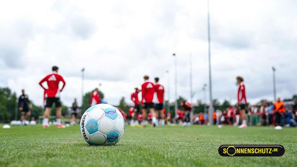 Ein Fußball liegt auf dem Rasen, während Spieler in roten Trikots im Hintergrund trainieren. Wolken bedecken den Himmel.