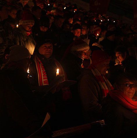 Menschen in Winterkleidung halten Kerzen in einer dunklen, festlichen Umgebung, wahrscheinlich bei einer Weihnachtsveranstaltung.