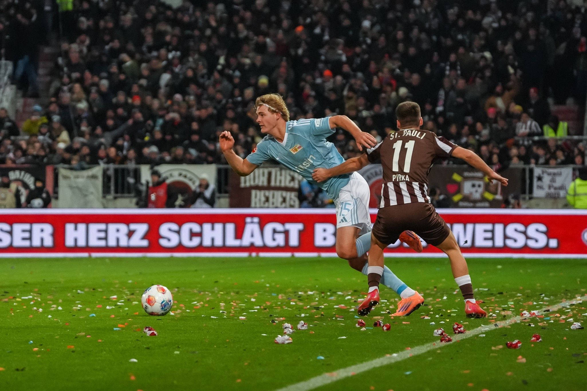 Ein Fußballspieler in blauer Uniform fällt, während ein Spieler in Braun ihn verfolgt. Der Platz ist mit Konfetti bedeckt.