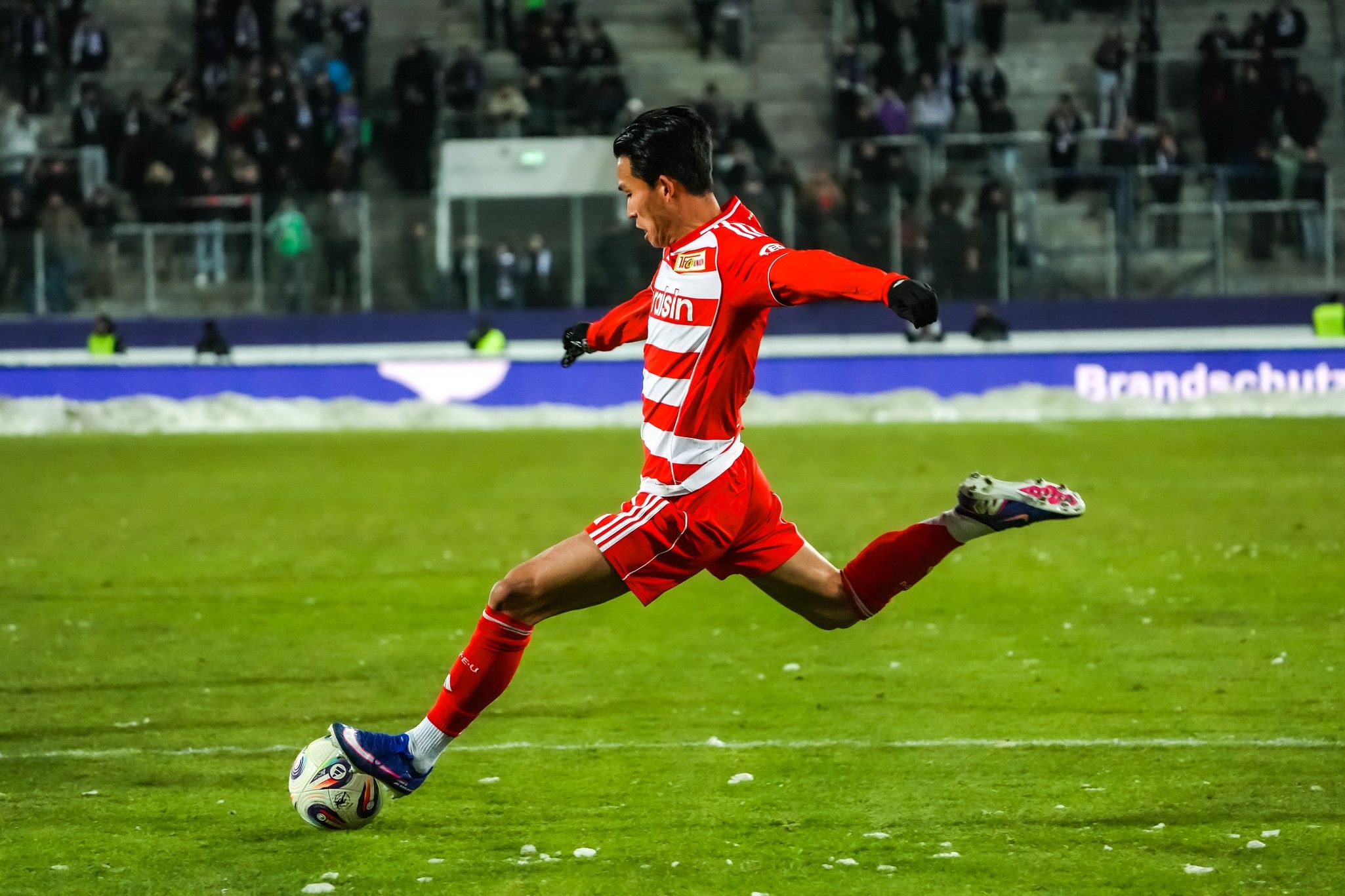 Ein Fußballspieler in rot-weißer Uniform tritt den Ball auf einem Rasenfeld, umgeben von jubelnden Zuschauern.