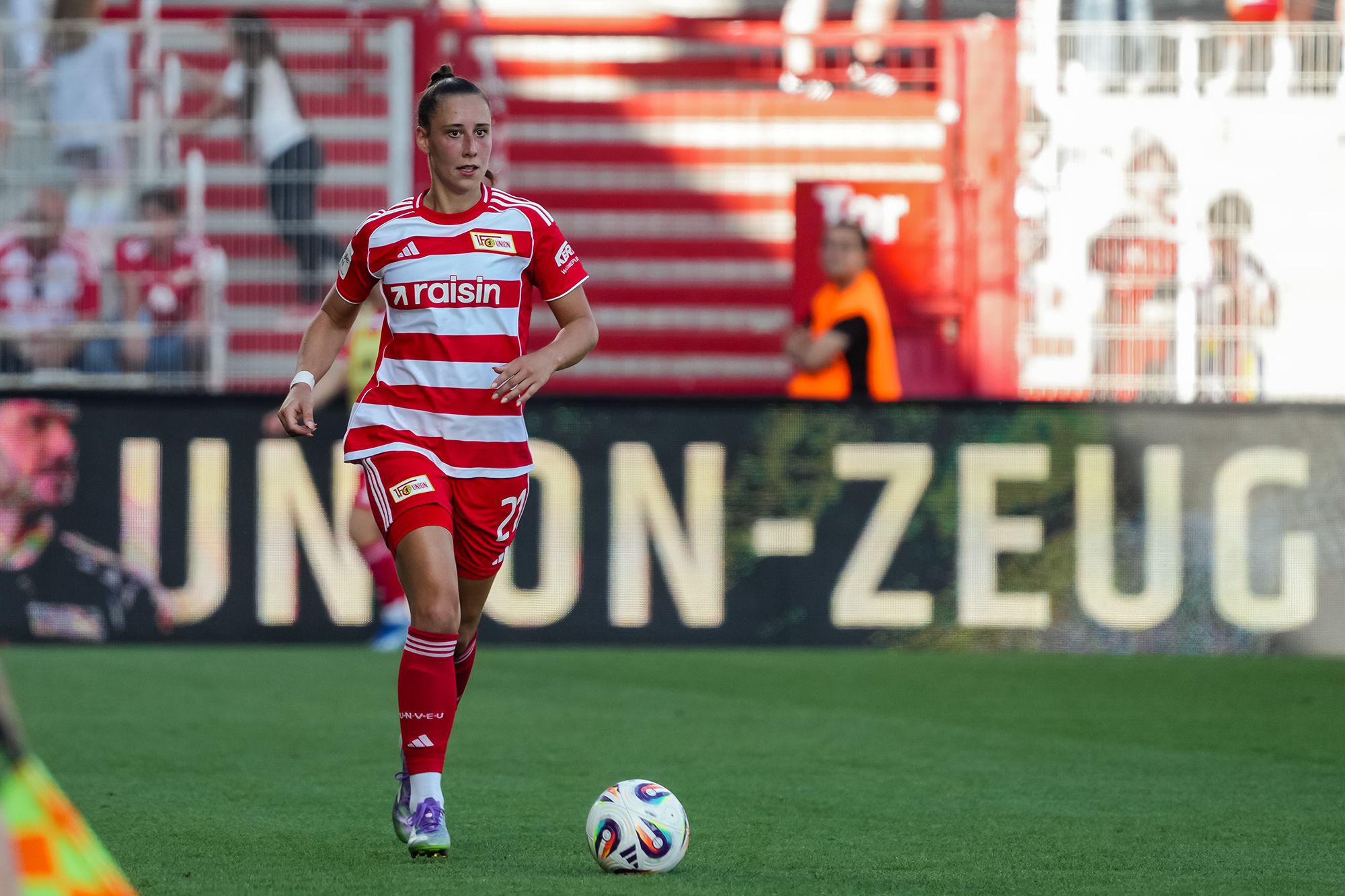 Spielerin in rot-weißer Vereinskleidung dribbelt den Ball auf dem Platz, im Hintergrund Fans und Werbung zu sehen.