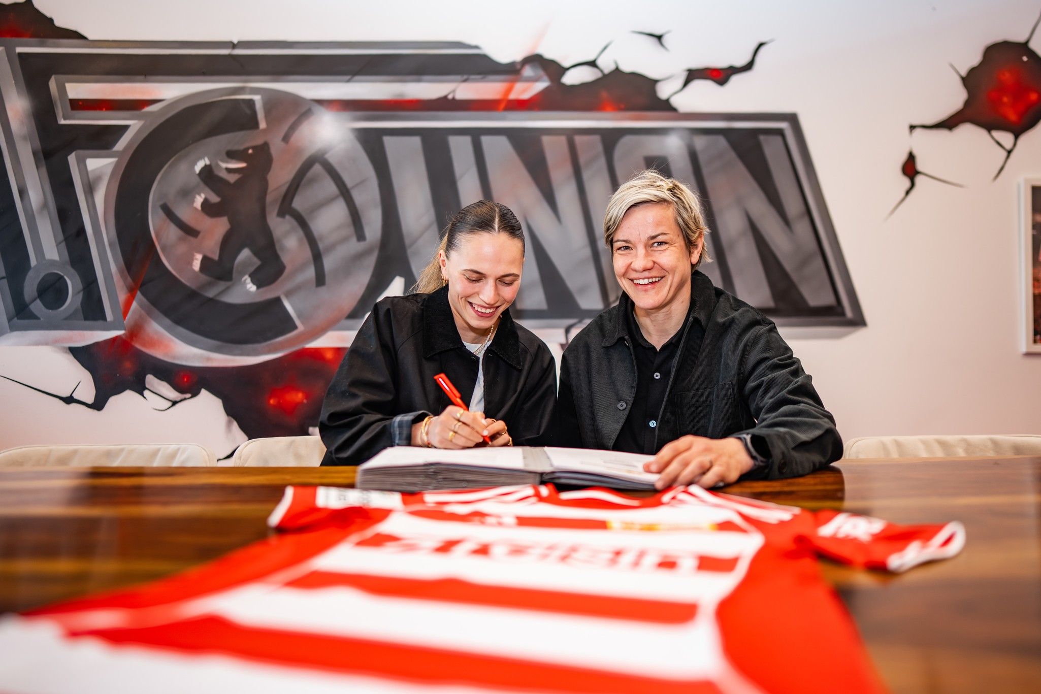 Zwei Frauen sitzen an einem Tisch und unterzeichnen Dokumente, während ein Fußballtrikot im Vordergrund liegt. Im Hintergrund ist ein Logo sichtbar.