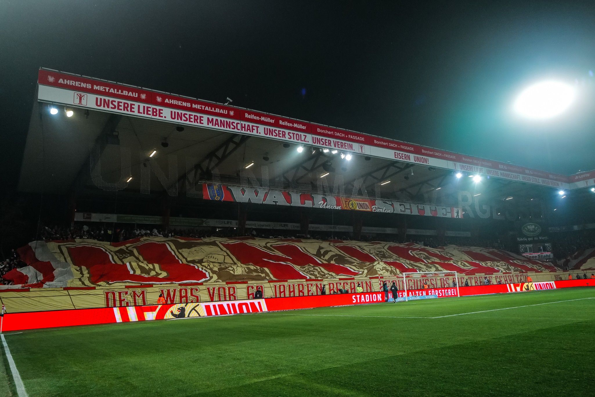 Fankurve mit großem Banner in Rot und Weiß, beleuchtet, die Stadionatmosphäre während eines Spiels zeigt.