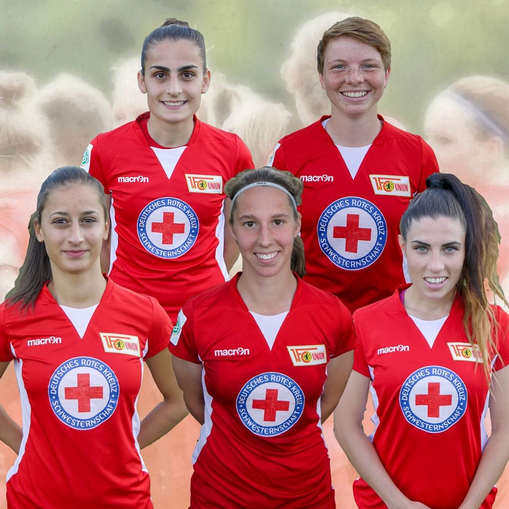 Fünf Fußballspielerinnen in roten Trikots mit einem Kreuz-Symbol, lächeln in die Kamera. Unscharfer Hintergrund.
