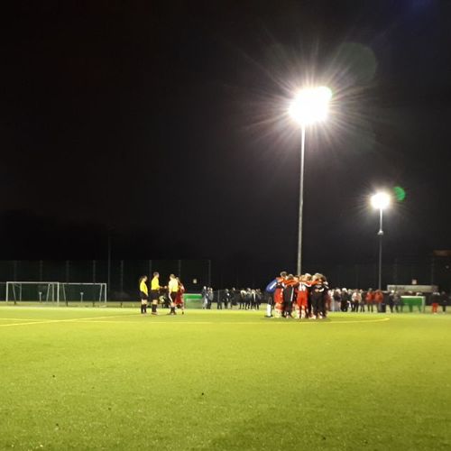 Spielende auf einem Fußballplatz bei Nacht, mit beleuchteten Spielfeldern und Spielern in roten und schwarzen Trikots.