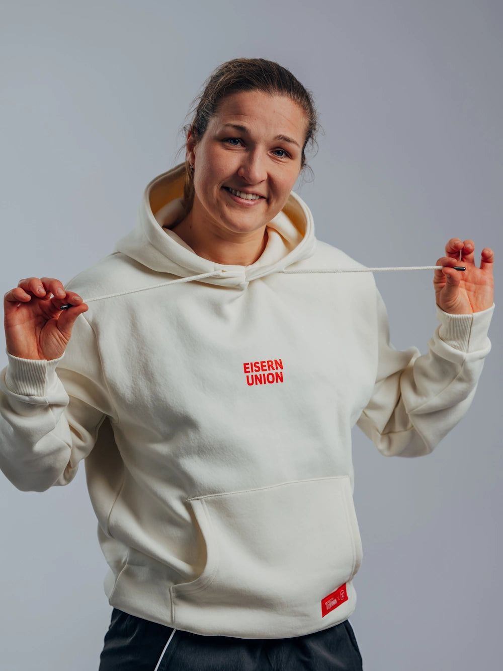 Eine Person in einem weißen Hoodie mit dem Aufdruck "EISERN UNION" hält den Hoodie-Zug in den Händen und lächelt.