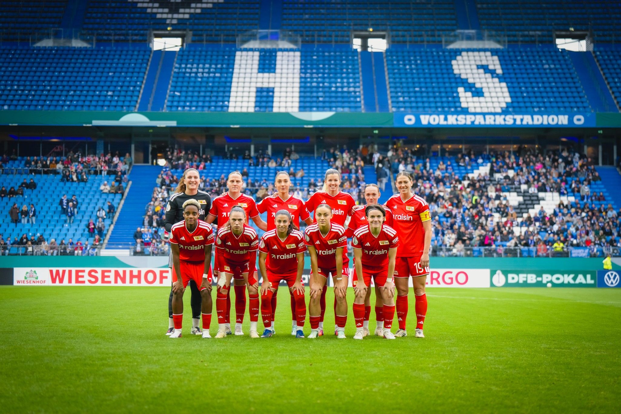 Frauenfußballmannschaft in roten Trikots posiert auf dem Spielfeld im Volksparkstadion. Zuschauer im Hintergrund.