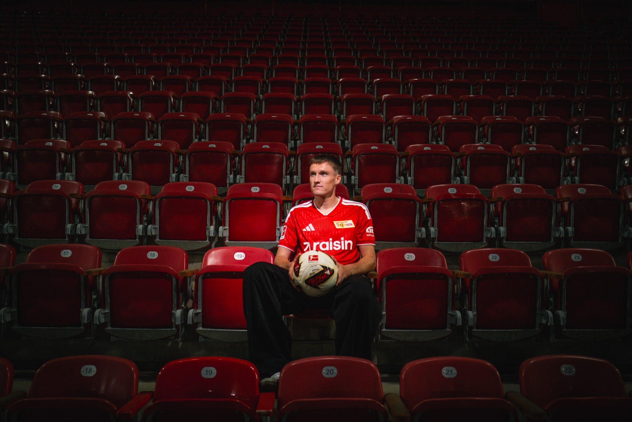 Ein Spieler in einem roten Trikot sitzt allein auf einem Stuhl in einem leeren Stadion mit roten Sitzreihen.