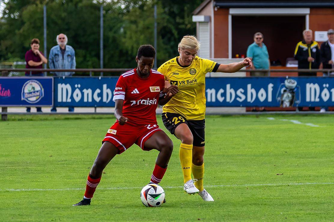 Zwei Fußballspieler im Wettkampf um den Ball auf einem grünen Platz, Zuschauer im Hintergrund.
