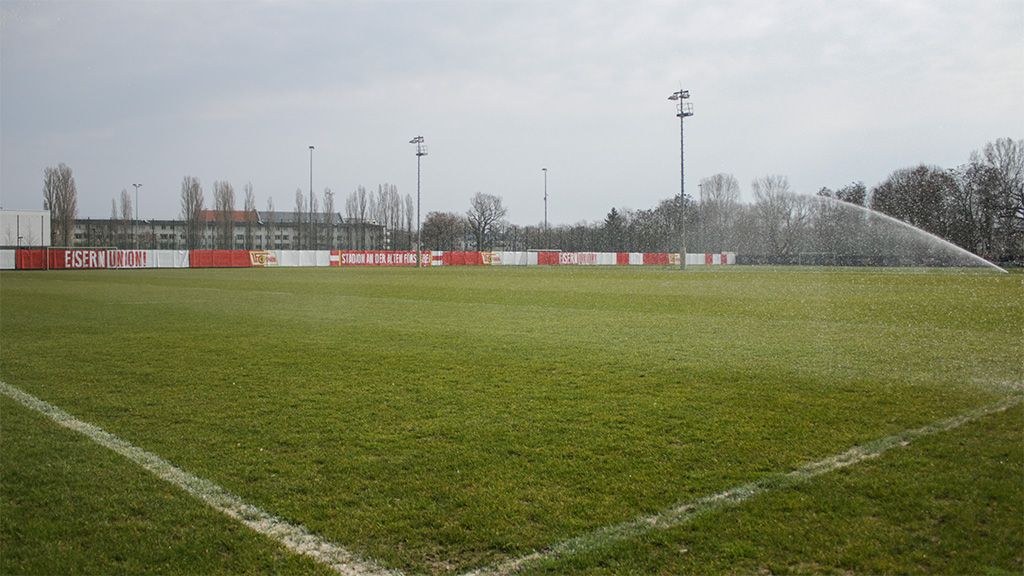 Rasenplatz mit Sprinkleranlage, grüner Rasen, Umrandung in rot und weiß, auf einem Sportgelände unter bewölktem Himmel.