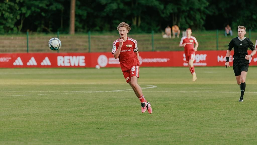 Ein junger Spieler in rotem Fußballtrikot springt, um den Ball auf dem Spielfeld zu erreichen. Im Hintergrund sind weitere Spieler und Zuschauer.