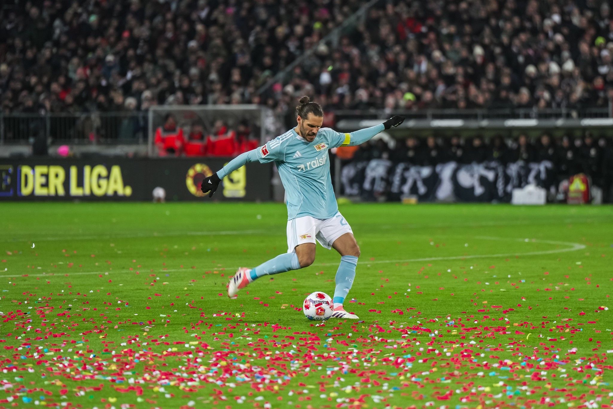 Ein Fußballspieler in blauer Uniform schießt einen Ball auf einem Rasenfeld, das mit buntem Konfetti bedeckt ist.