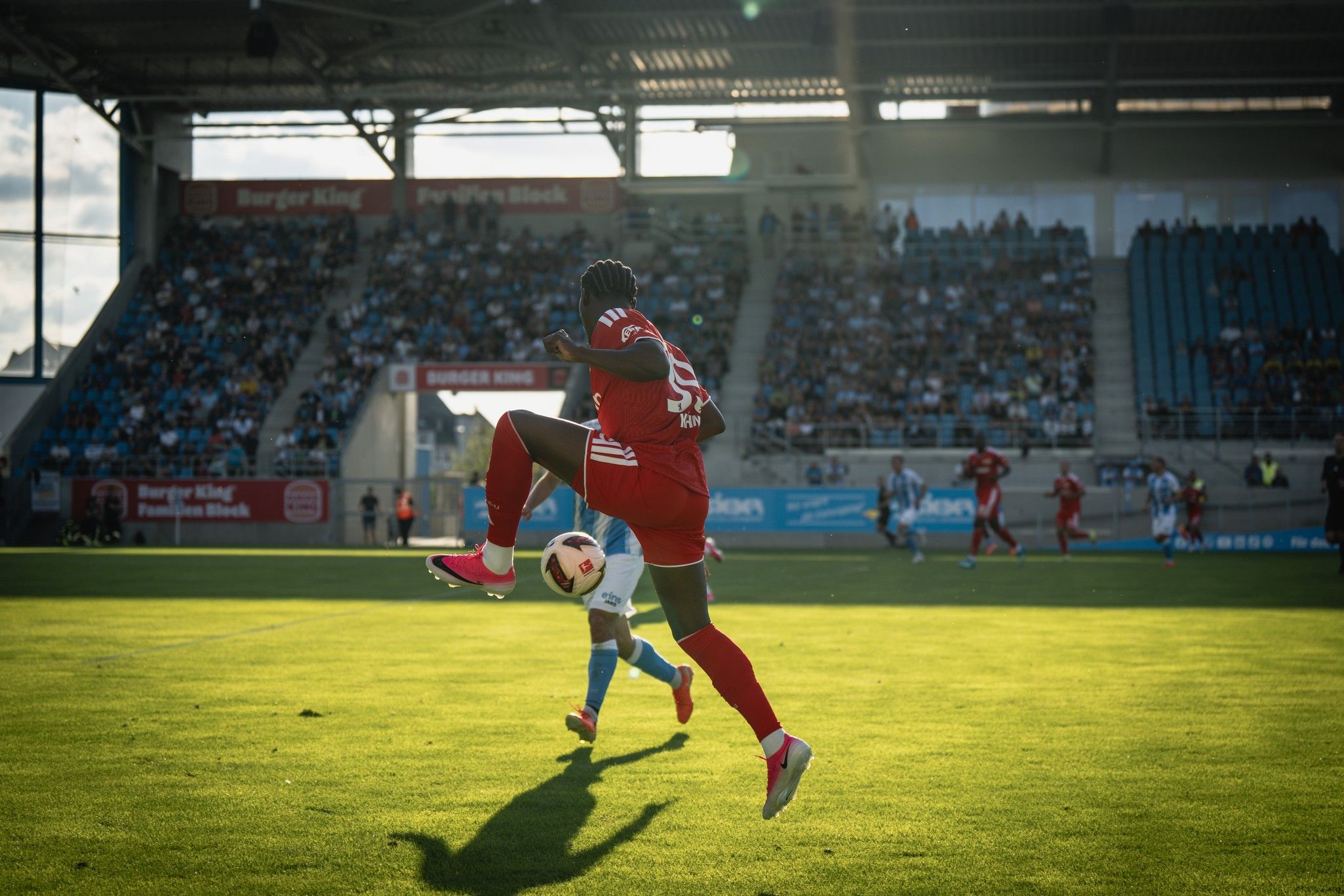 Ein Fußballspieler in rotem Trikot springt über einen Gegner auf einem gut gepflegten Rasenfeld, mit Zuschauern im Hintergrund.