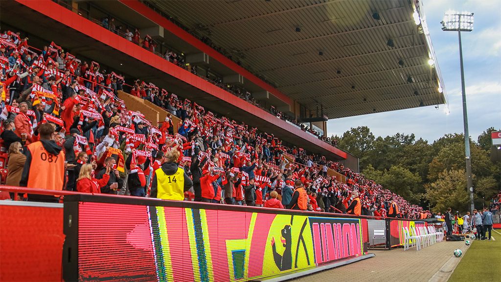 Fans in roten T-Shirts schwenken Schals in einem Stadion. Die Tribüne ist gut gefüllt und die Stimmung ist festlich.