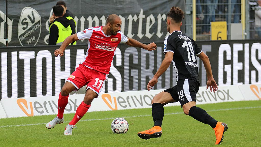 Spielszene eines Fußballmatches mit Spielern in roten und schwarzen Trikots, die um den Ball kämpfen.
