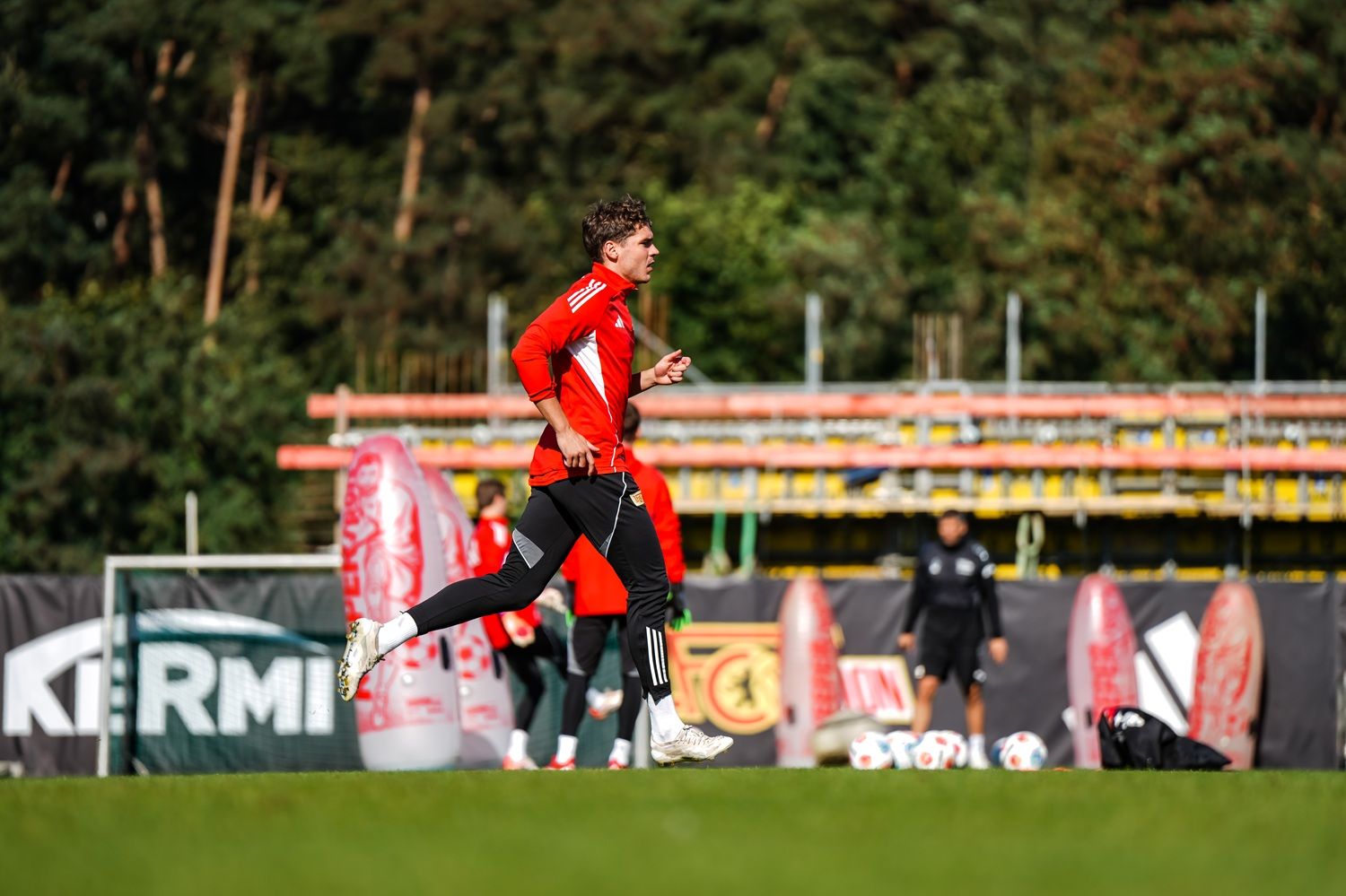 Ein Fußballspieler in rotem Trikot läuft auf einem Trainingsplatz, umgeben von weiteren Spielern und Bäumen im Hintergrund.