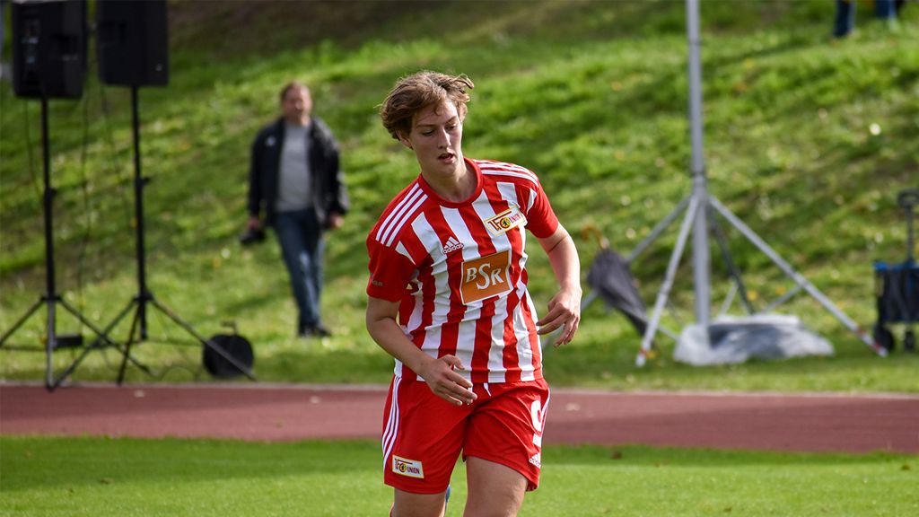 Ein junger Fußballspieler in rot-weiß gestreiftem Trikot läuft auf einem Rasenplatz. Im Hintergrund sind Zuschauer zu sehen.