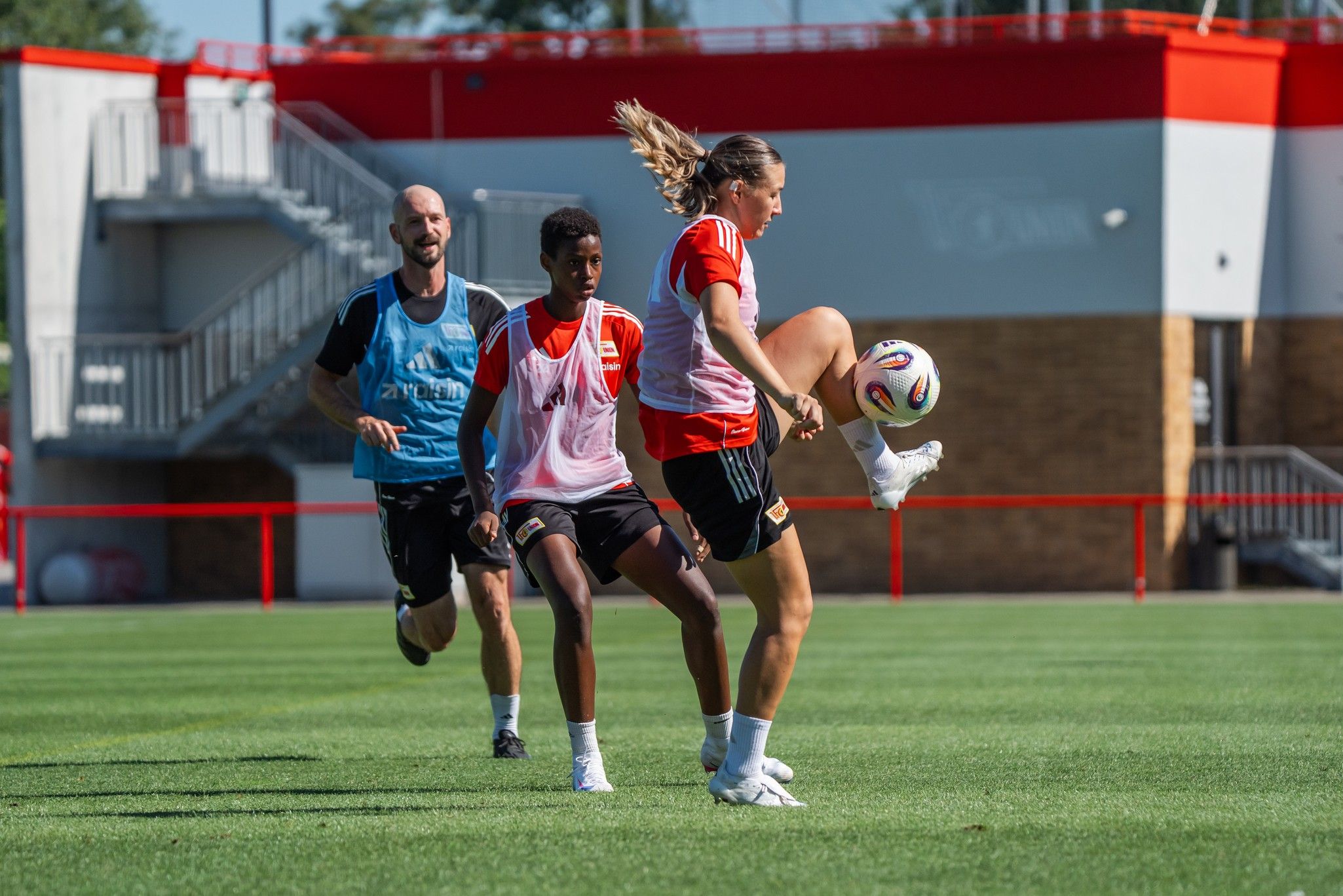 Drei Personen trainieren auf einem Fußballplatz; eine Spielerin dribbelt den Ball, während die anderen zuschauen.