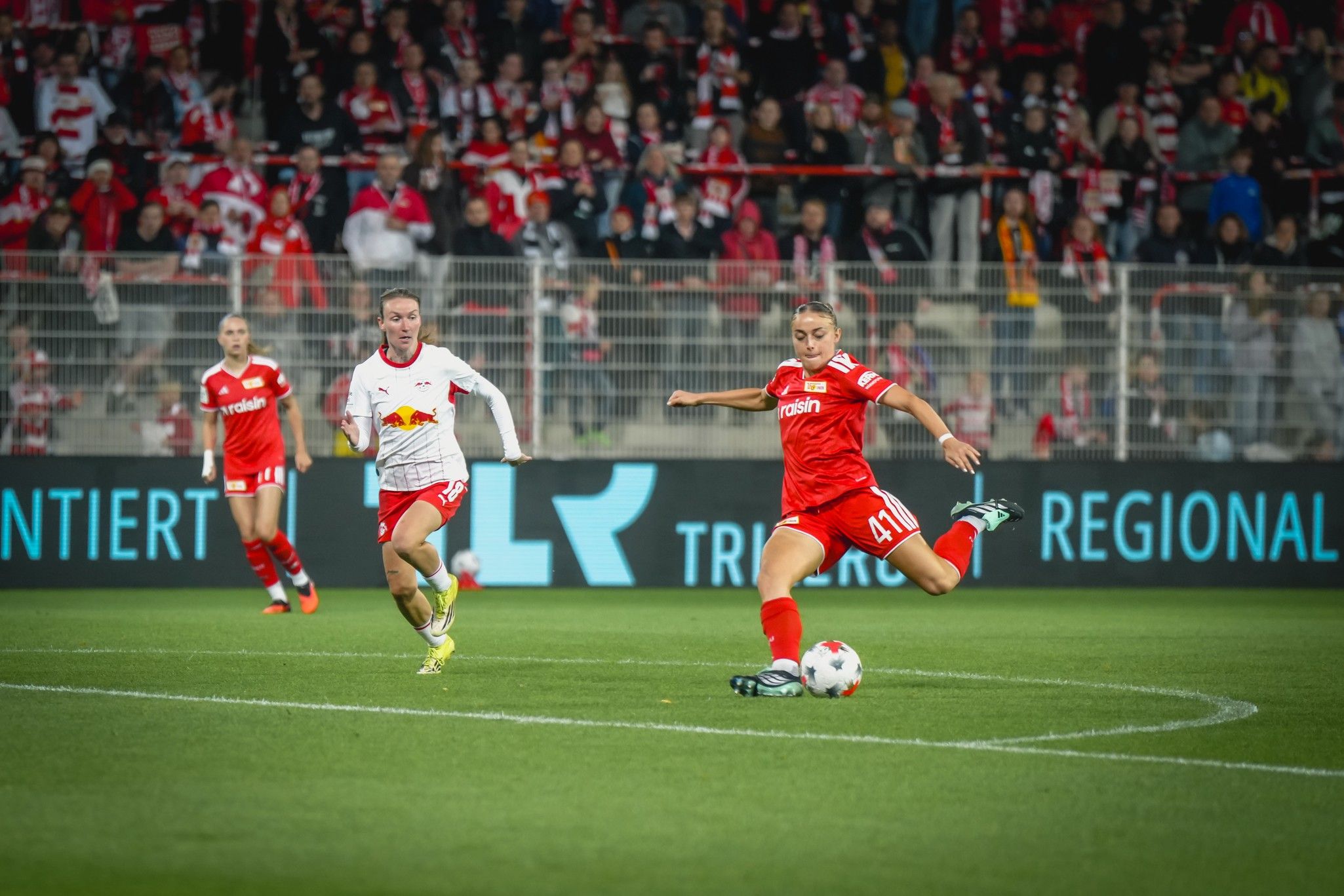 Ein Spieler im roten Trikot schießt den Ball, während ein Gegenspieler im weißen Trikot ihn verfolgt. Stadionatmosphäre im Hintergrund.