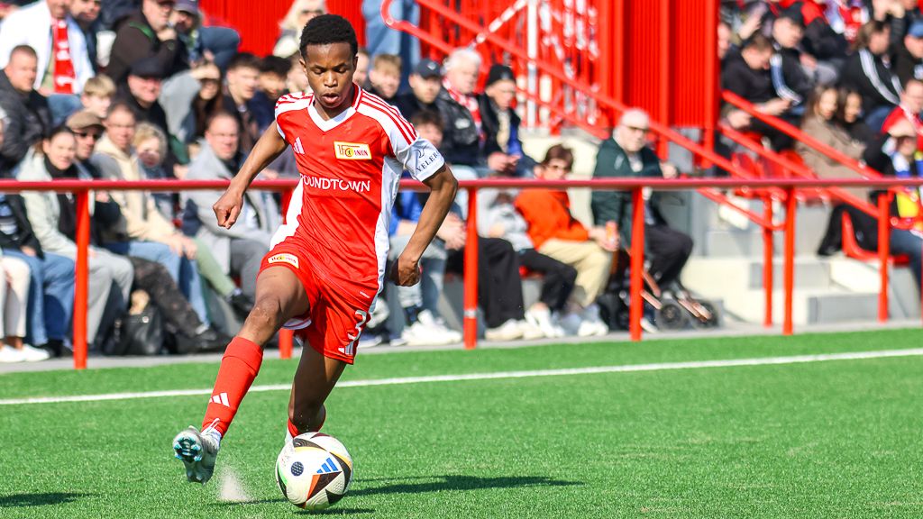 Ein junger Fußballspieler in rotem Trikot zieht mit dem Ball auf einem Rasenplatz, umgeben von jubelnden Zuschauern.