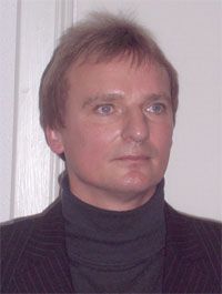 Porträt eines Mannes mit kurzem, blondem Haar, der einen dunklen Rollkragenpullover und einen gestreiften Anzug trägt, vor einer weißen Wand.