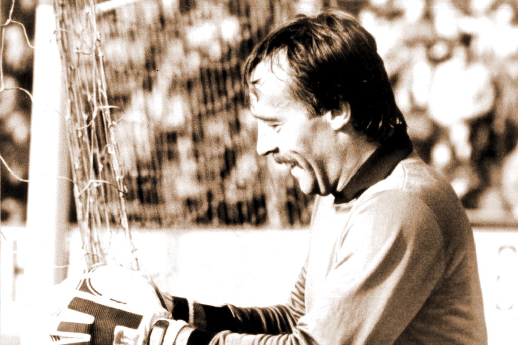 A goalkeeper in a football game, smiling while standing in front of the goal and holding his gloves. Sepia tone.