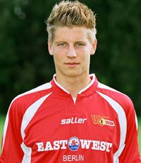 Junger Mann in rotem Fußballtrikot vor einem unscharfen Hintergrund, mit kurzen, blonden Haaren und blauen Augen.