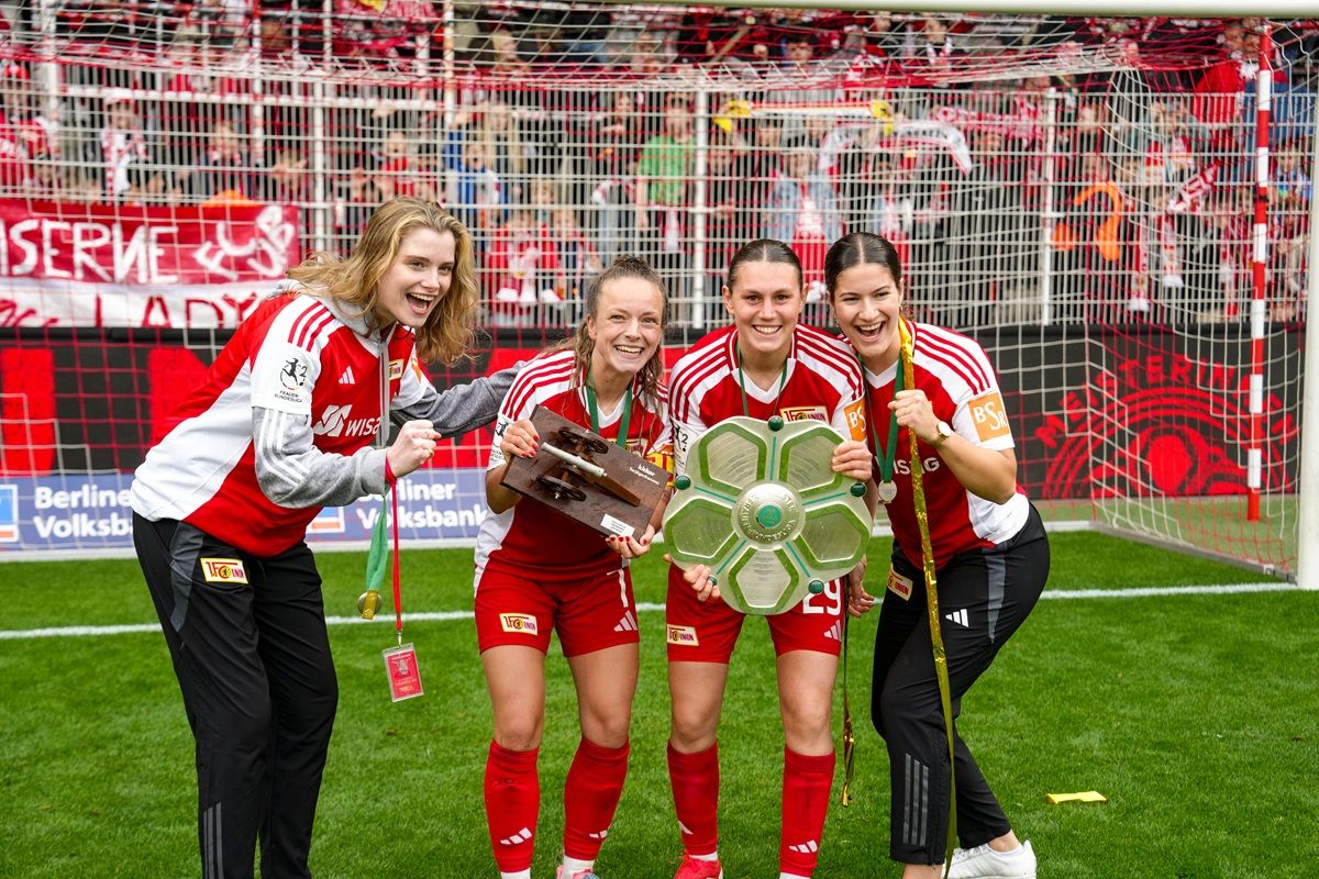 Vier Fußballspielerinnen in roten Trikots präsentieren stolz ihre Trophäen auf dem Spielfeld vor einer jubelnden Kulisse.