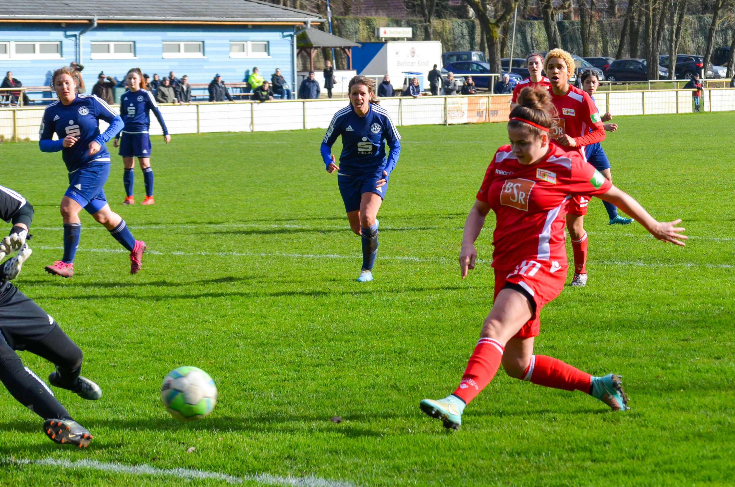 Eine Spielerin in rotem Trikot schießt auf das Tor, während Spielerinnen in blauen Trikots im Hintergrund auf den Ball reagieren.