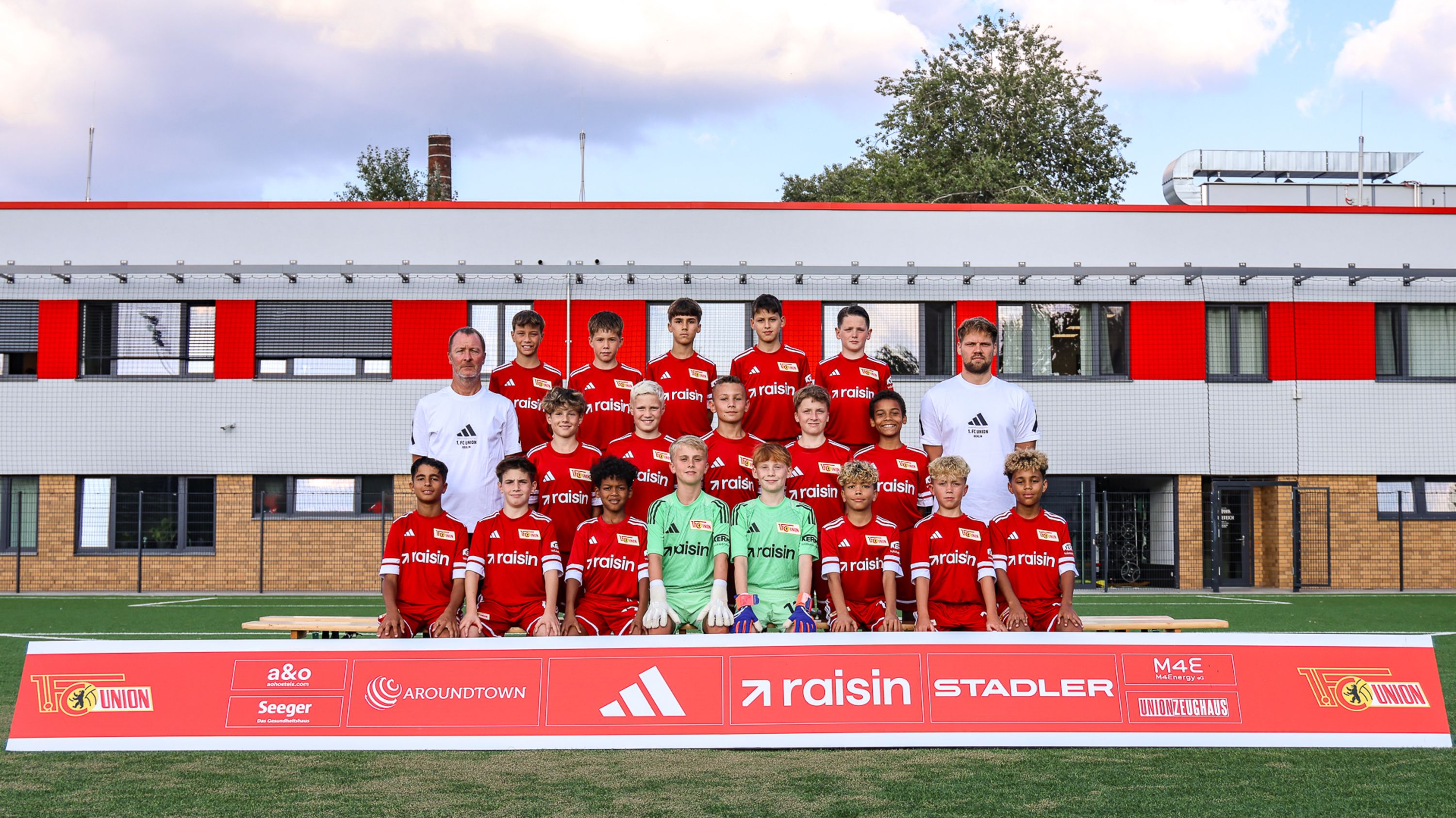 Jugendfußballmannschaft in roten Trikots steht vor einem Gebäude, mit Trainern und Sponsorenlogo auf einem Banner.