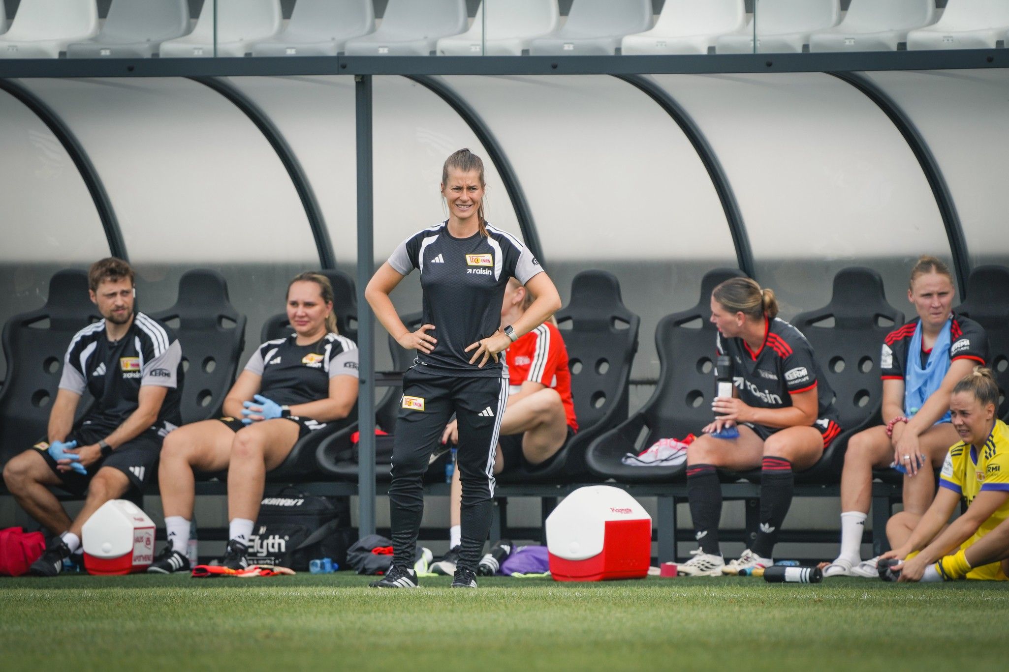 Eine Trainerin steht an der Seitenlinie, während Spielerinnen auf der Ersatzbank sitzen, bei einem Fußballtraining.