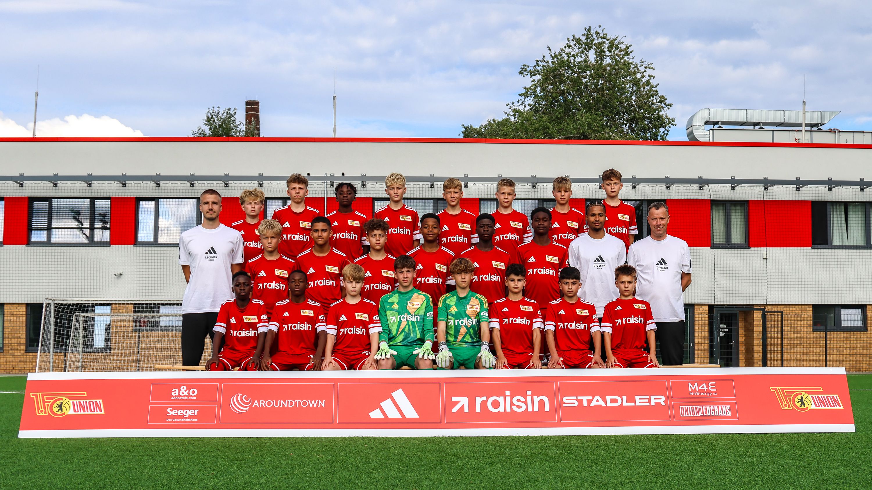 Mannschaftsbild einer Jugendfußballmannschaft in roten Trikots, posierend vor einem Sportgelände mit Sponsorenbanner.