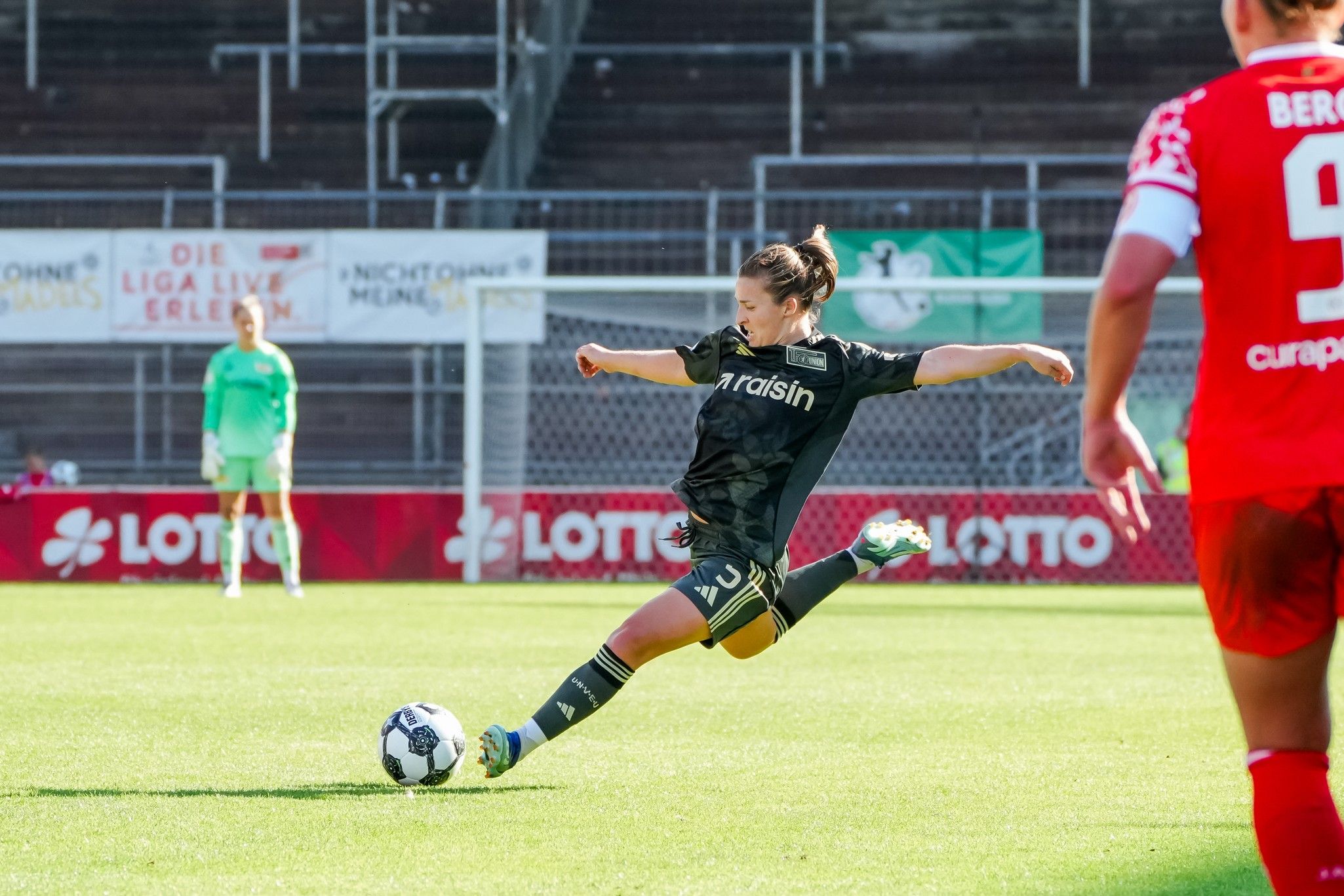 Eine Fußballspielerin in schwarzer Trikotausrüstung tritt einen Ball auf dem Spielfeld. Im Hintergrund sind Spieler und ein Tor zu sehen.