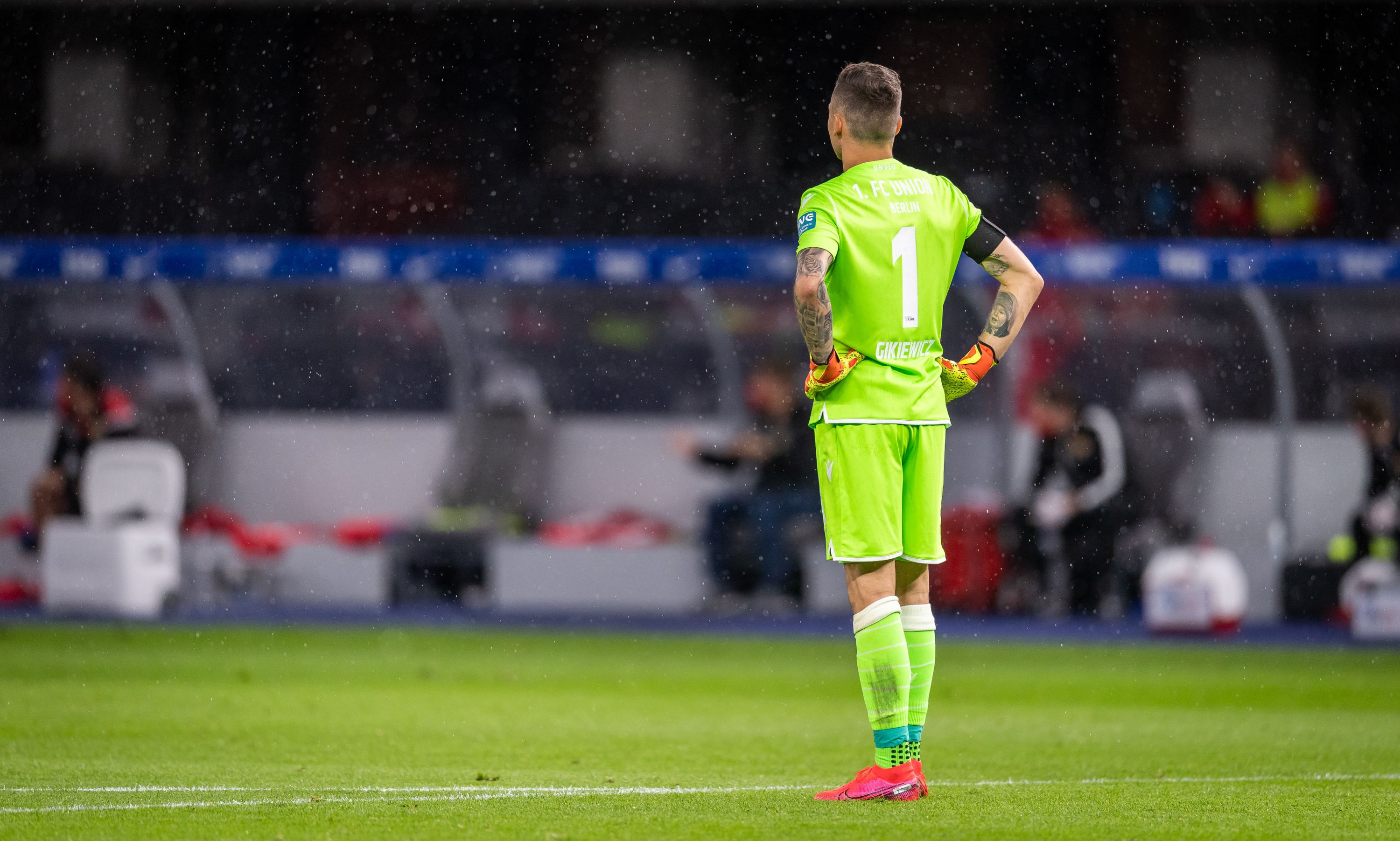 A goalkeeper in a green jersey stands motionless in the rain at the sideline of a soccer field.