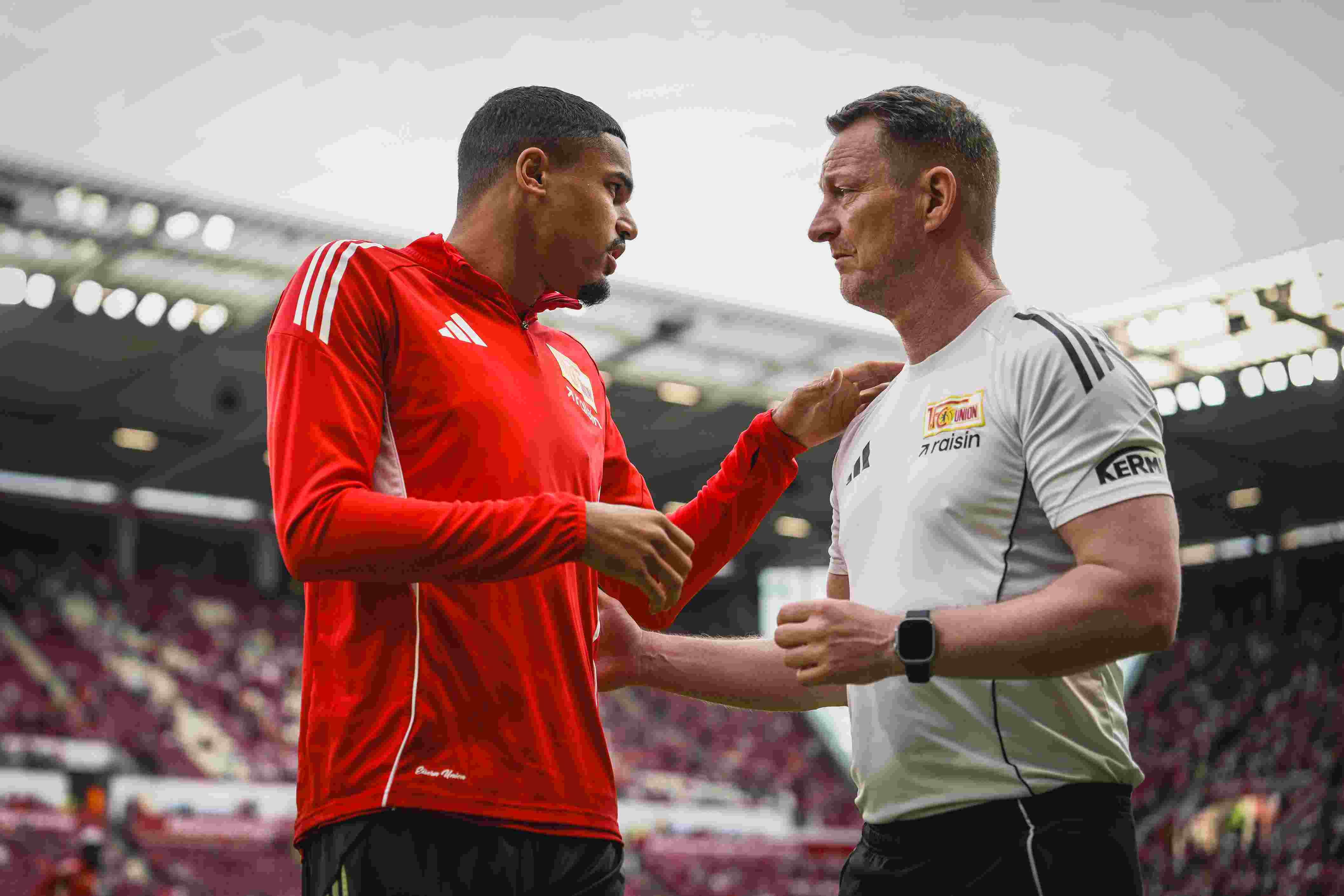 Zwei Männer stehen auf einem Fußballplatz, einer trägt ein rotes Trikot, der andere ein Schiedsrichter-Outfit, beide im Gespräch.