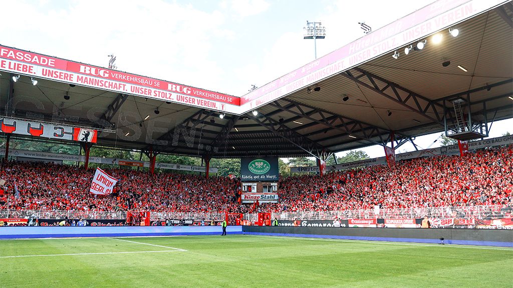 Das Bild zeigt ein voll besetztes Fußballstadion mit leidenschaftlichen Fans und einem grünen Spielfeld.