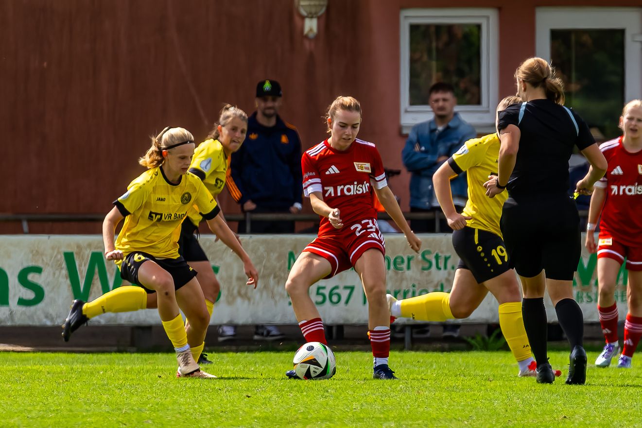 Eine Fußballspielerin mit rotem Trikot dribbelt den Ball, während ihre Gegnerinnen in gelben Trikots sie herausfordern.
