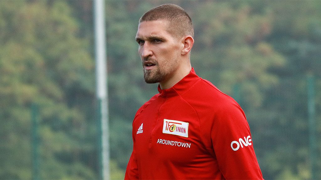 A soccer player in a red jersey stands on the field, looking seriously into the distance. The background is blurred with trees.