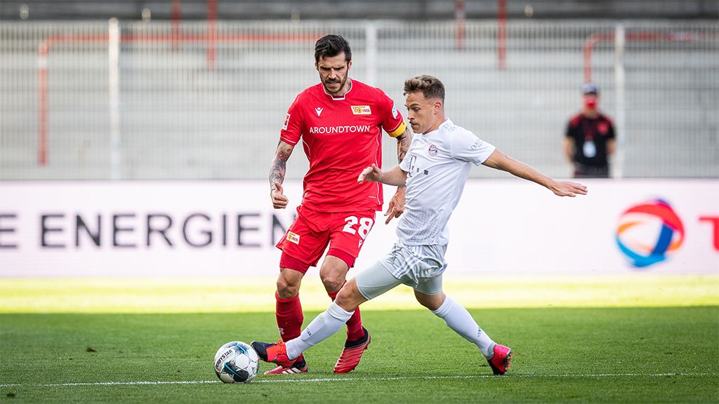 Zwei Fußballspieler kämpfen um den Ball auf einem Sportfeld, einer trägt ein rotes Trikot, der andere ein weißes Trikot.