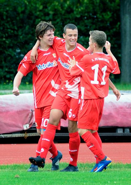 Drei Fußballspieler in roten Trikots feiern einen Treffer auf dem Spielfeld.