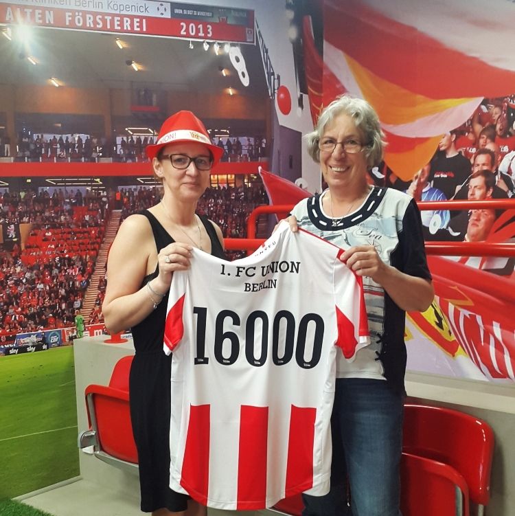 Zwei Frauen halten ein Trikot des 1. FC Union Berlin mit der Nummer 16000 vor einem Stadion-Hintergrund.