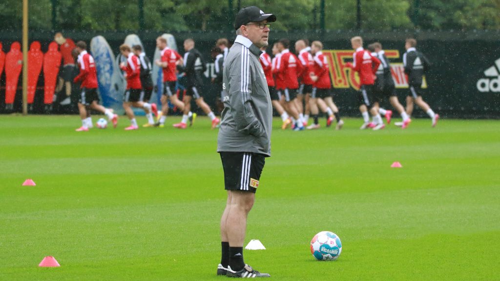 Ein Trainer steht an der Seitenlinie eines Fußballtrainingsgeländes, während Spieler im Hintergrund trainieren.